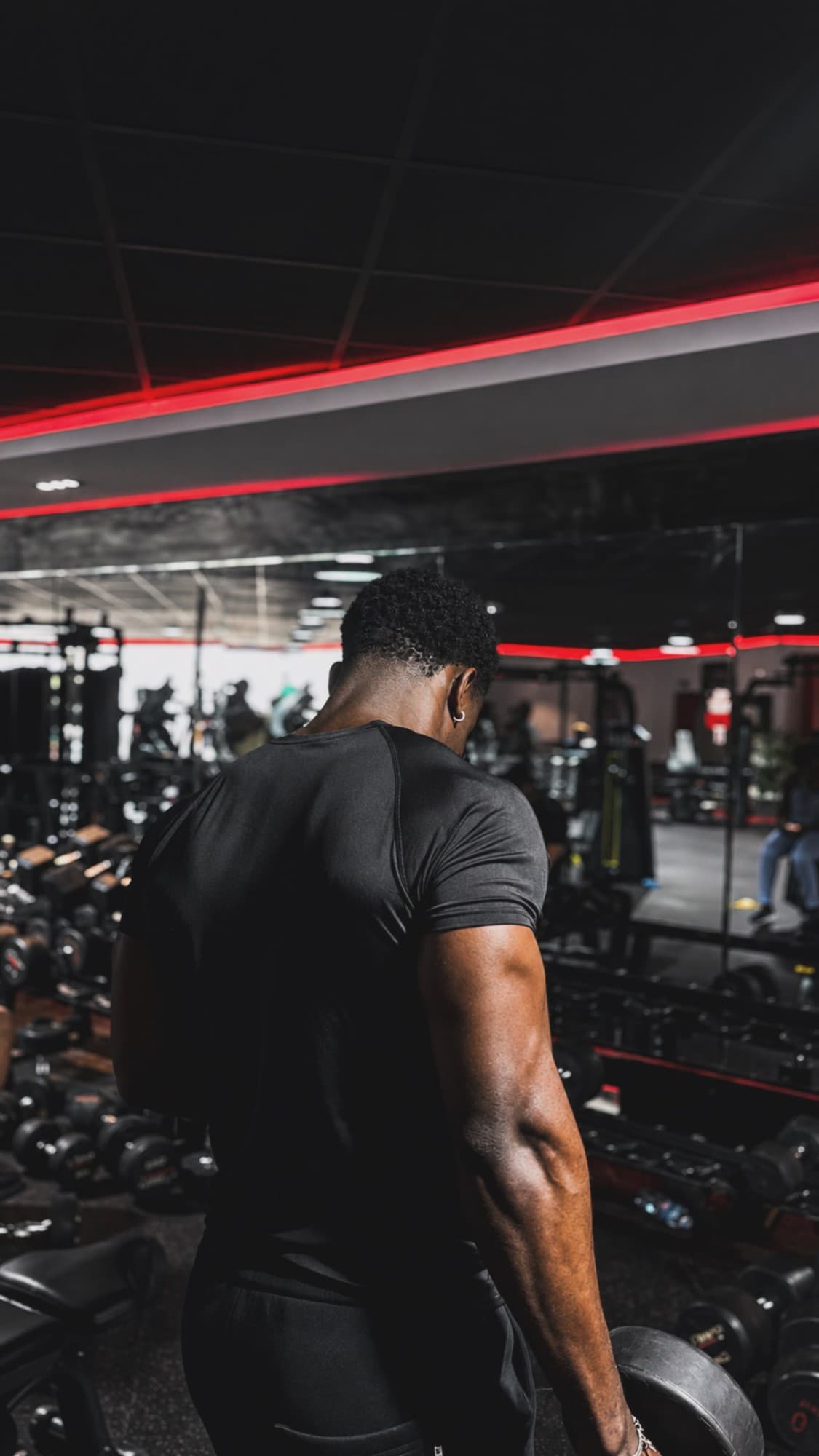 Tife in the black compression tee, seen from behind, holding a dumbbell in the gym